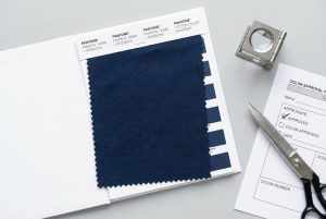Navy blue fabric swatch being compared against a Pantone color guide with an approval form and textile loupe for strict quality control.