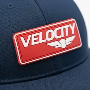 A macro photograph of a red and white 3D PVC "VELOCITY" logo patch on a navy blue perforated athletic performance cap, showing sharp relief and no cracking.